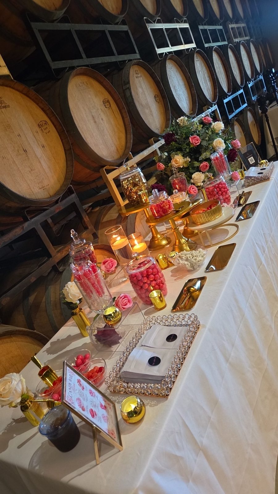 Candy bar table styled with gold stands and florals against wine barrels